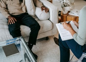 From above crop African American male in casual clothes sitting on comfy couch and answering professional female consultant questions in modern psychotherapy office